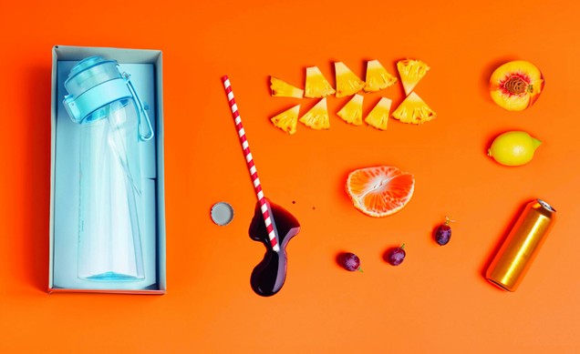 Flat lay: boxed blue bottle beside pineapple pieces, mandarin, grapes and a striped straw on an orange ground.