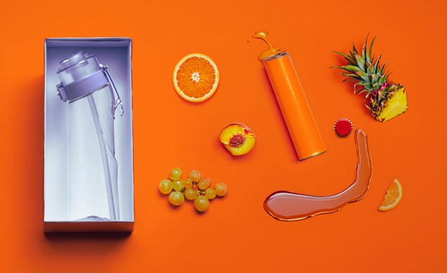 Flat lay: boxed clear bottle beside an orange slice, grapes, peach, pineapple and poured syrup on an orange ground.