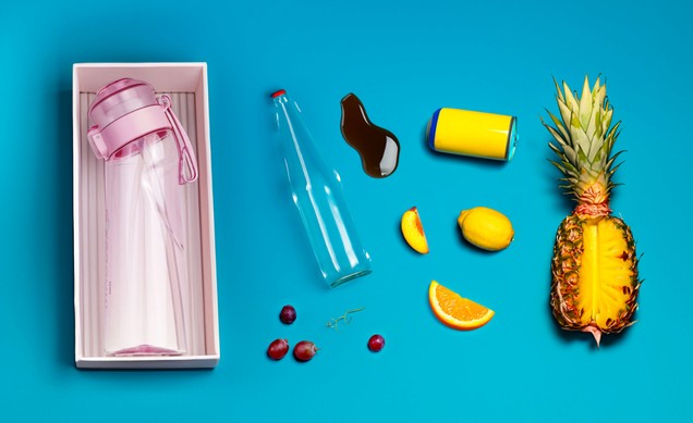 Flat lay: boxed pink bottle beside a glass bottle, a yellow can, lemons and halved pineapple on a blue ground.