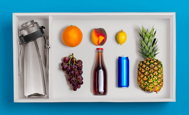 Flat lay: bottle set in a white tray beside whole fruit, a soda bottle and a can on a blue ground.